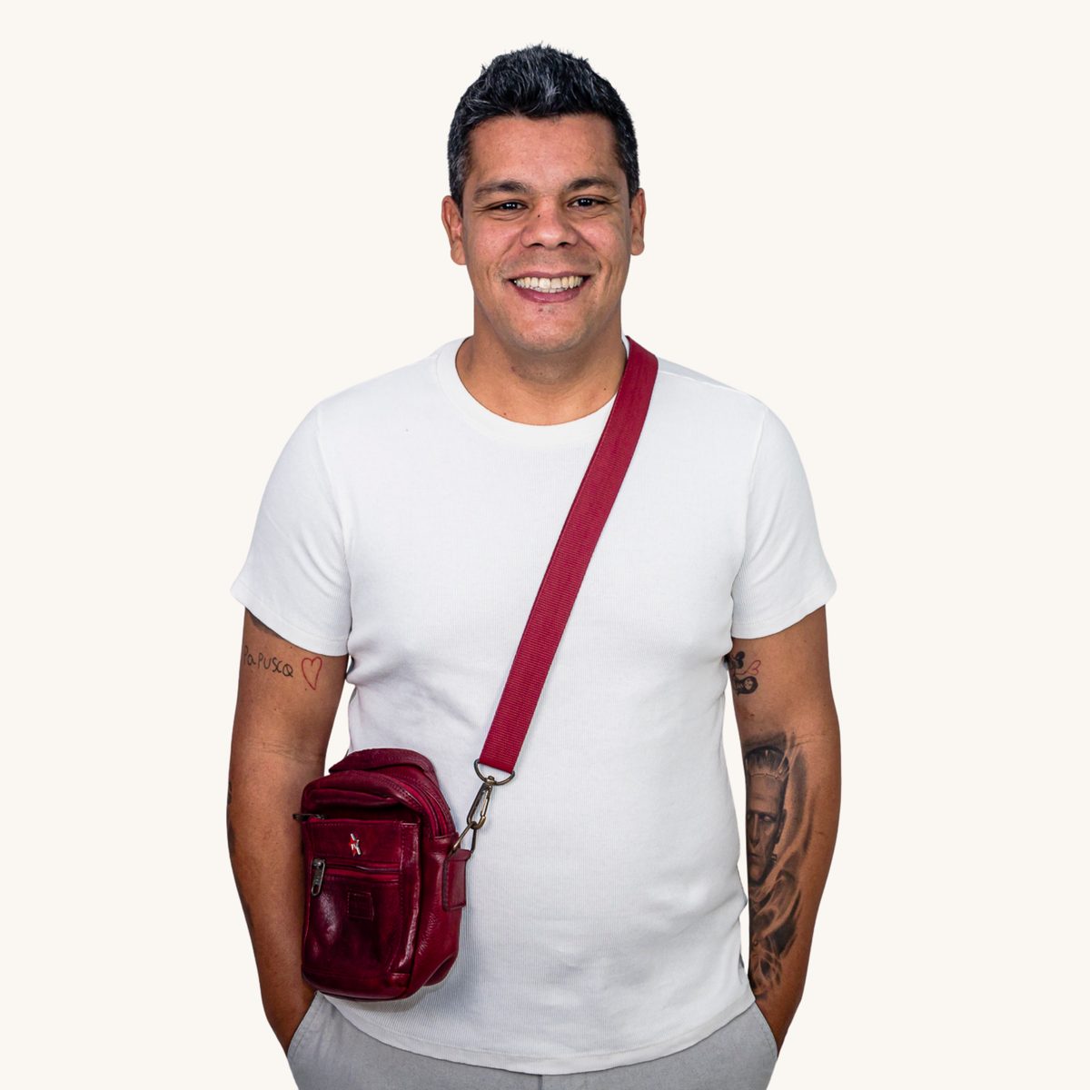 Retrato de Leonel Querino: homem negro, sorriso aberto, camiseta branca, com bolsa transversal vinho, em fundo claro.